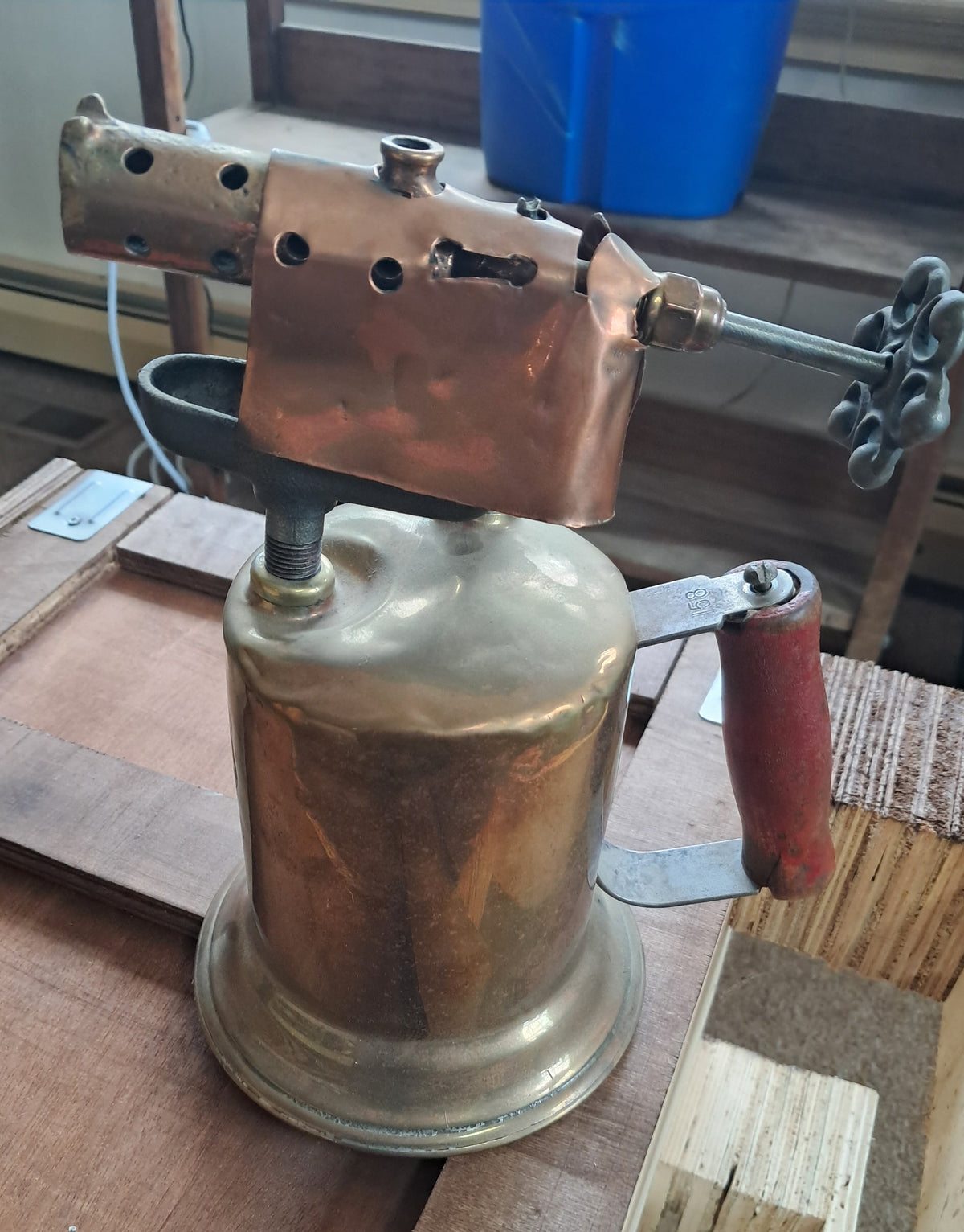 A vintage brass torch light with a copper body, brass burner head, and a red-painted handle, resting on a wooden workbench.